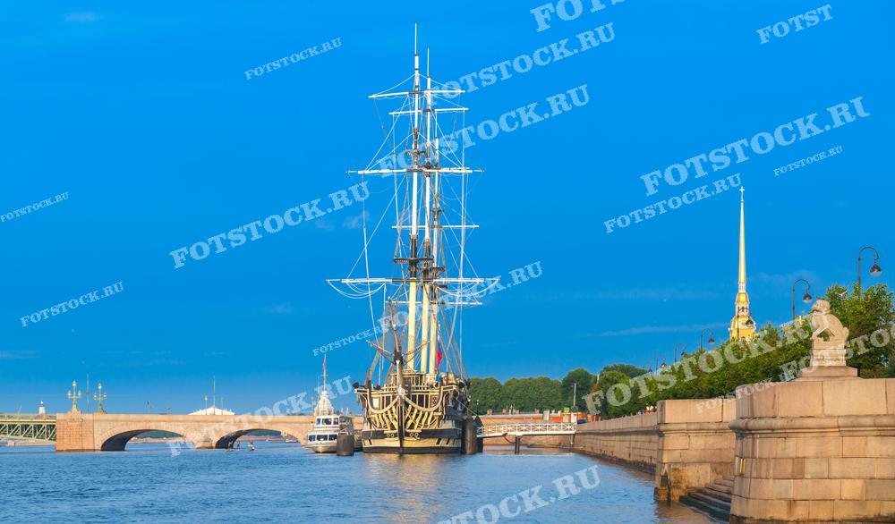 Фотография показывает ясный день в Санкт-Петербурге с видом на реку Нева и реконструированный парусник, который является частью морского музея. Вдалеке виден шпиль Петропавловской крепости. Это фото подойдёт для использования в туристических буклетах и постах в социальных сетях, рекламирующих достопримечательности города, а также может быть использовано в учебных материалах по истории мореплавания и культуре Санкт-Петербурга.